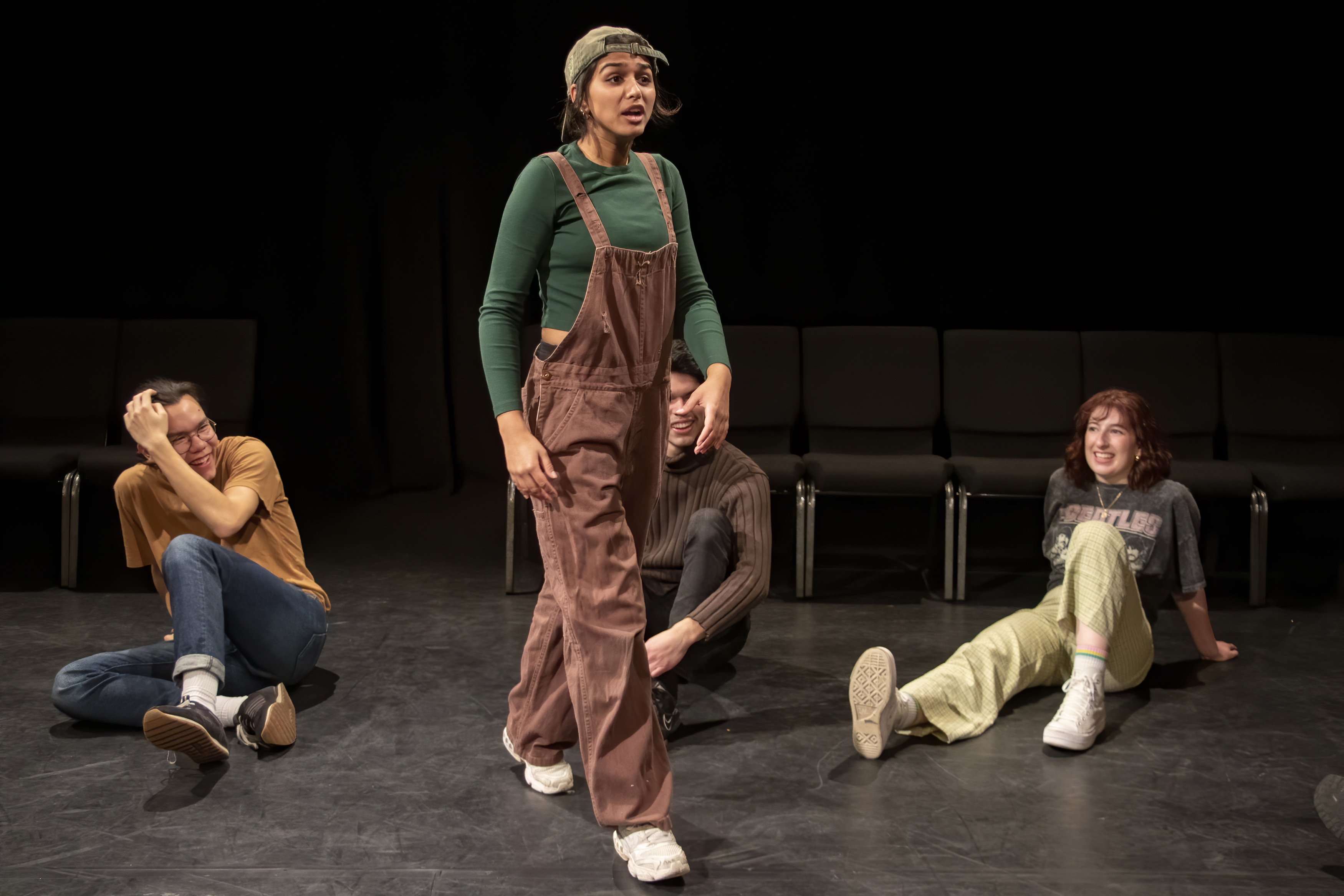 One student walking in mid-performance as other students look at her, seated on the floor of the black box theatre